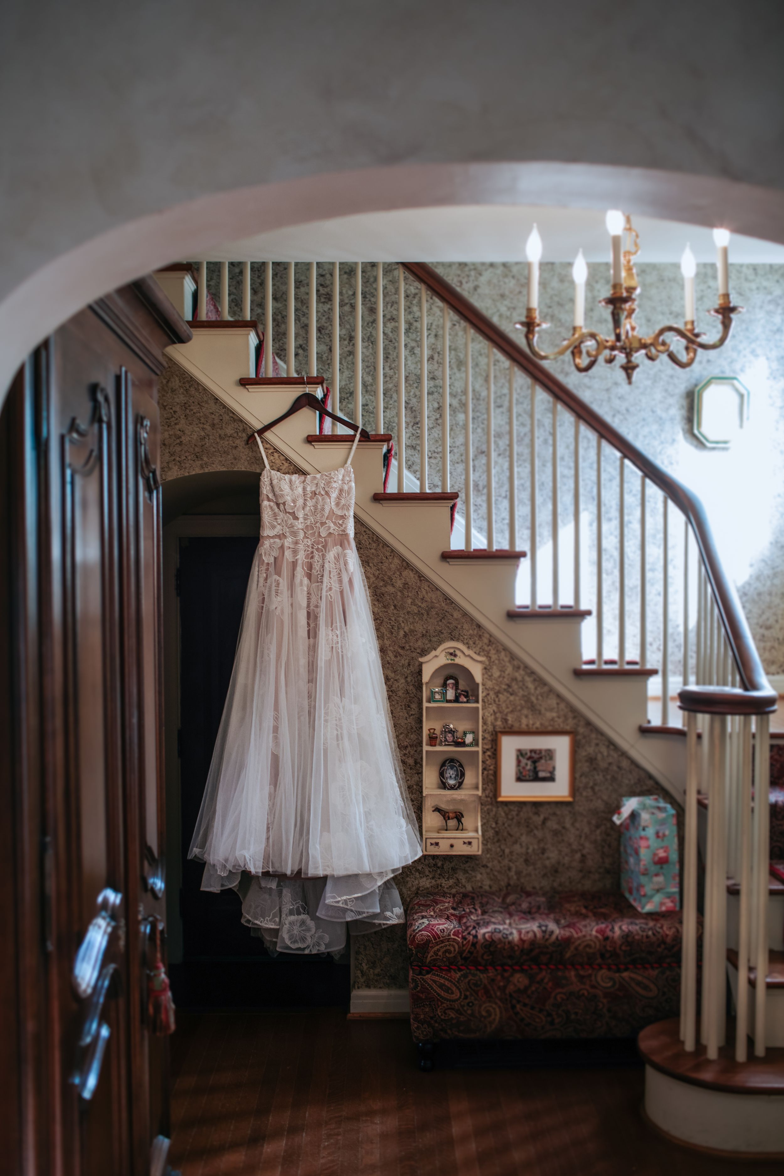 A delicate lace wedding dress hangs from a wooden staircase in a vintage-style home interior with wallpaper and chandelier.