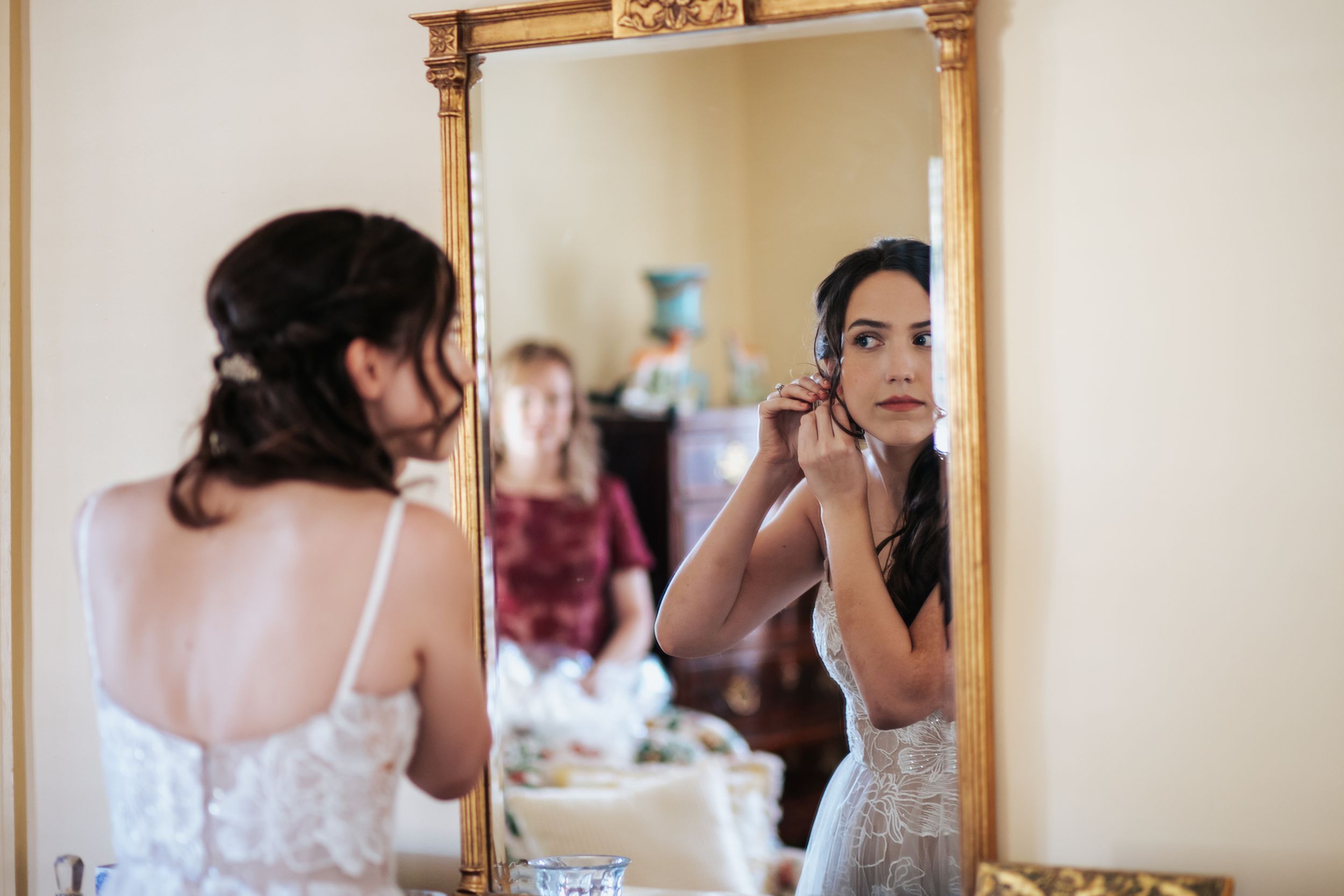 A reflection in an ornate gold mirror shows someone in a white lace dress getting ready for a special occasion.