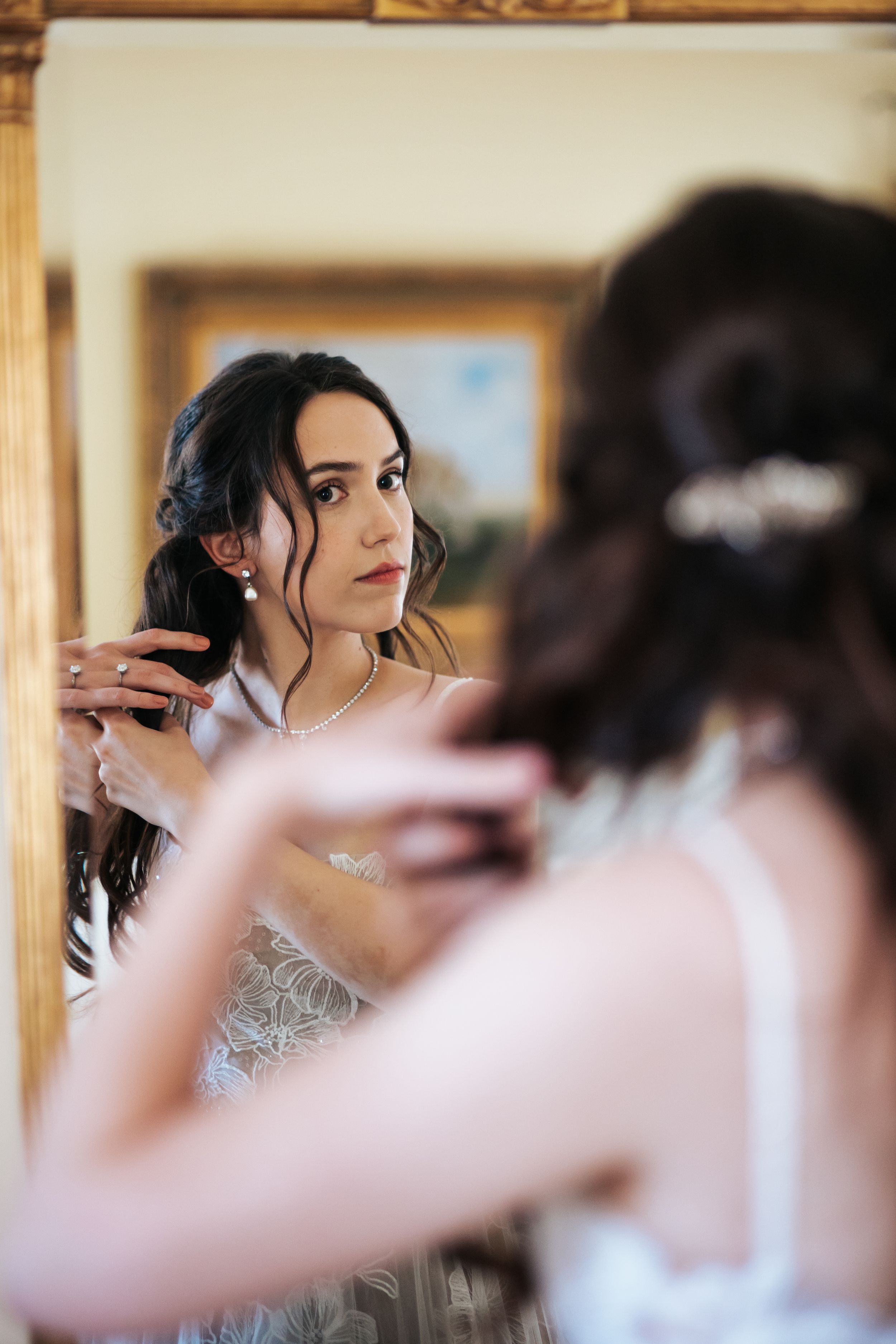 A bride gets ready in front of a mirror while her reflection shows her adjusting her hair.