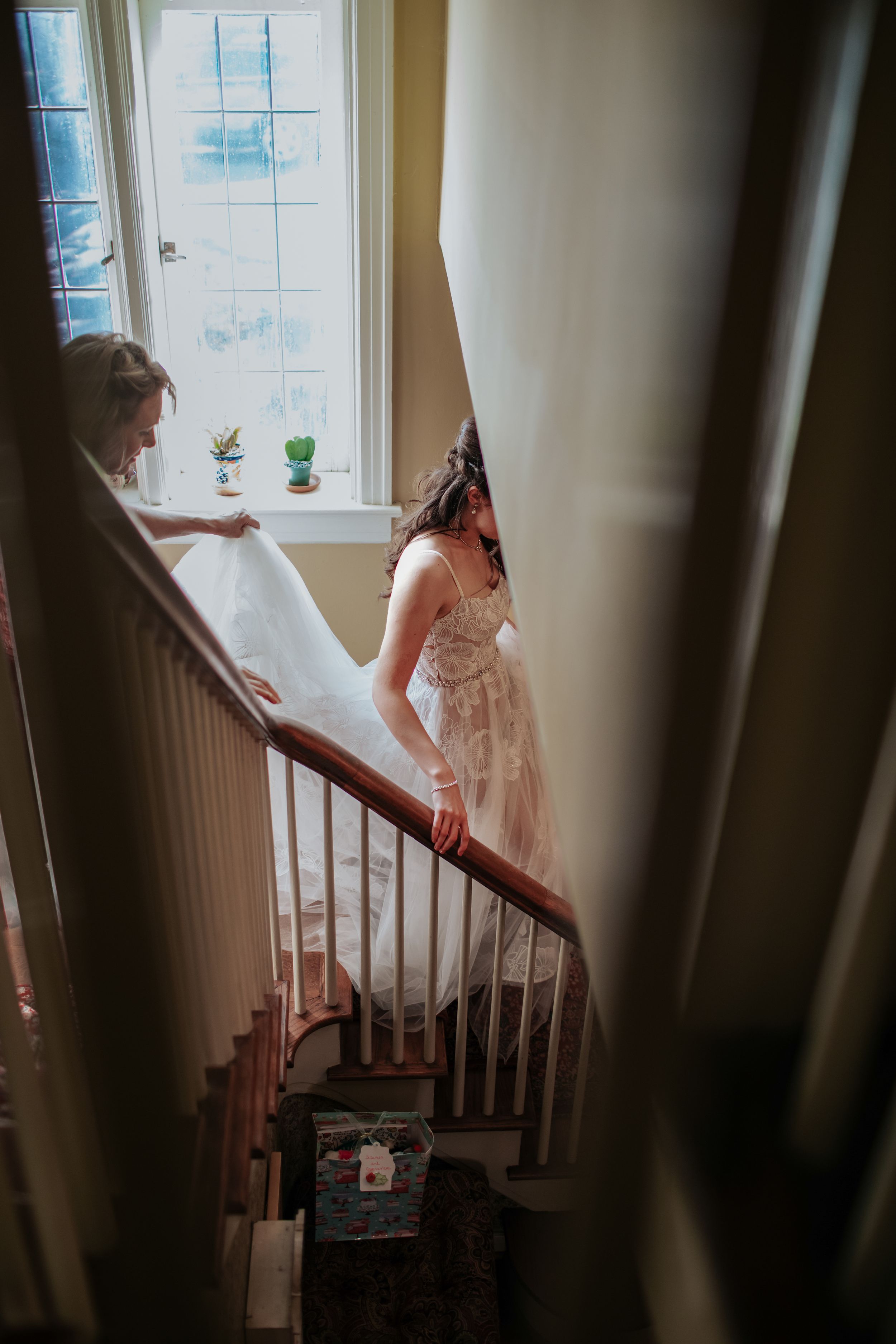 Bride in a flowing white wedding dress descends a sunlit staircase in an elegant home.