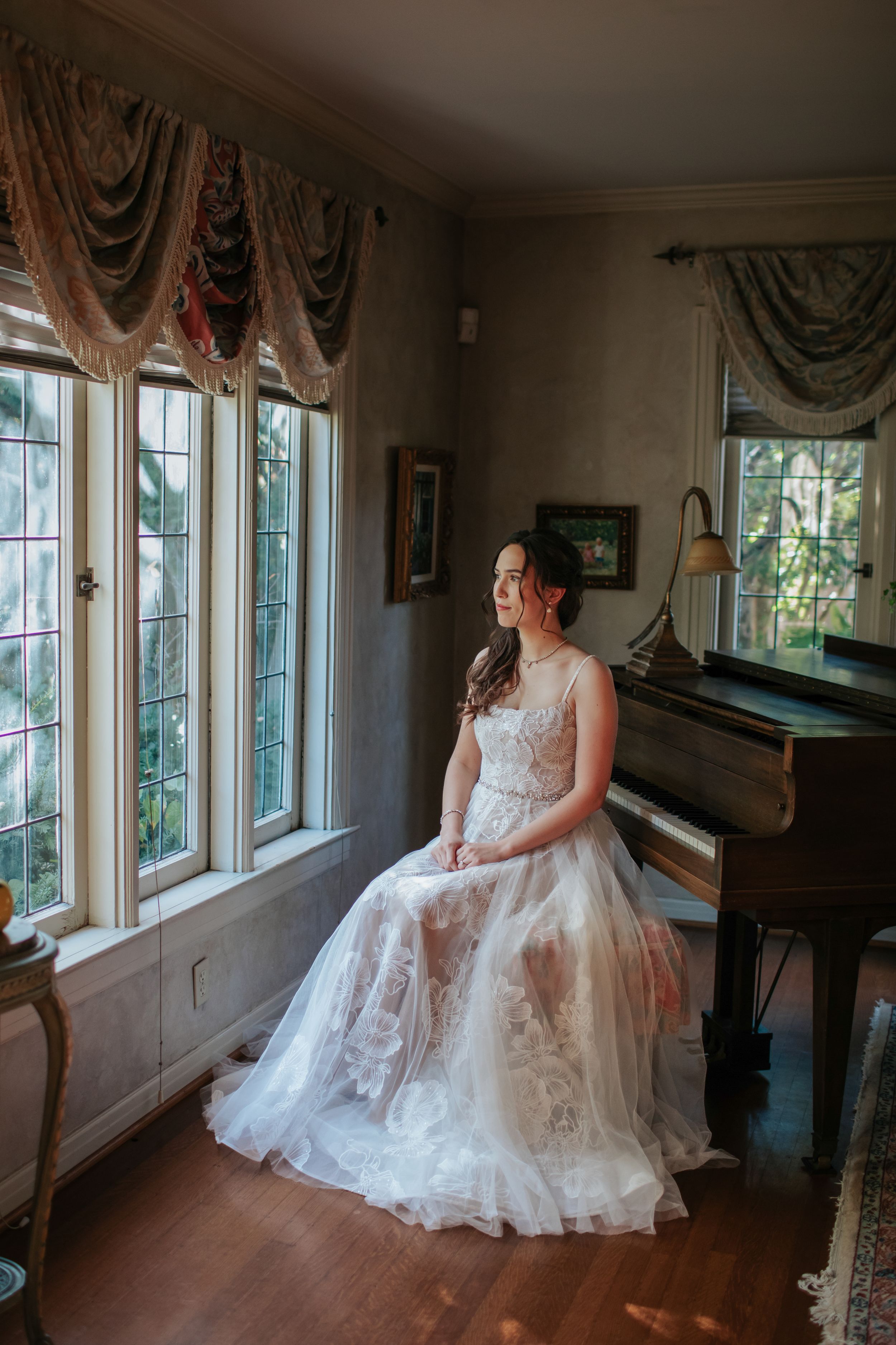 An elegant wedding dress is showcased in a sunlit room with a piano and draped windows.