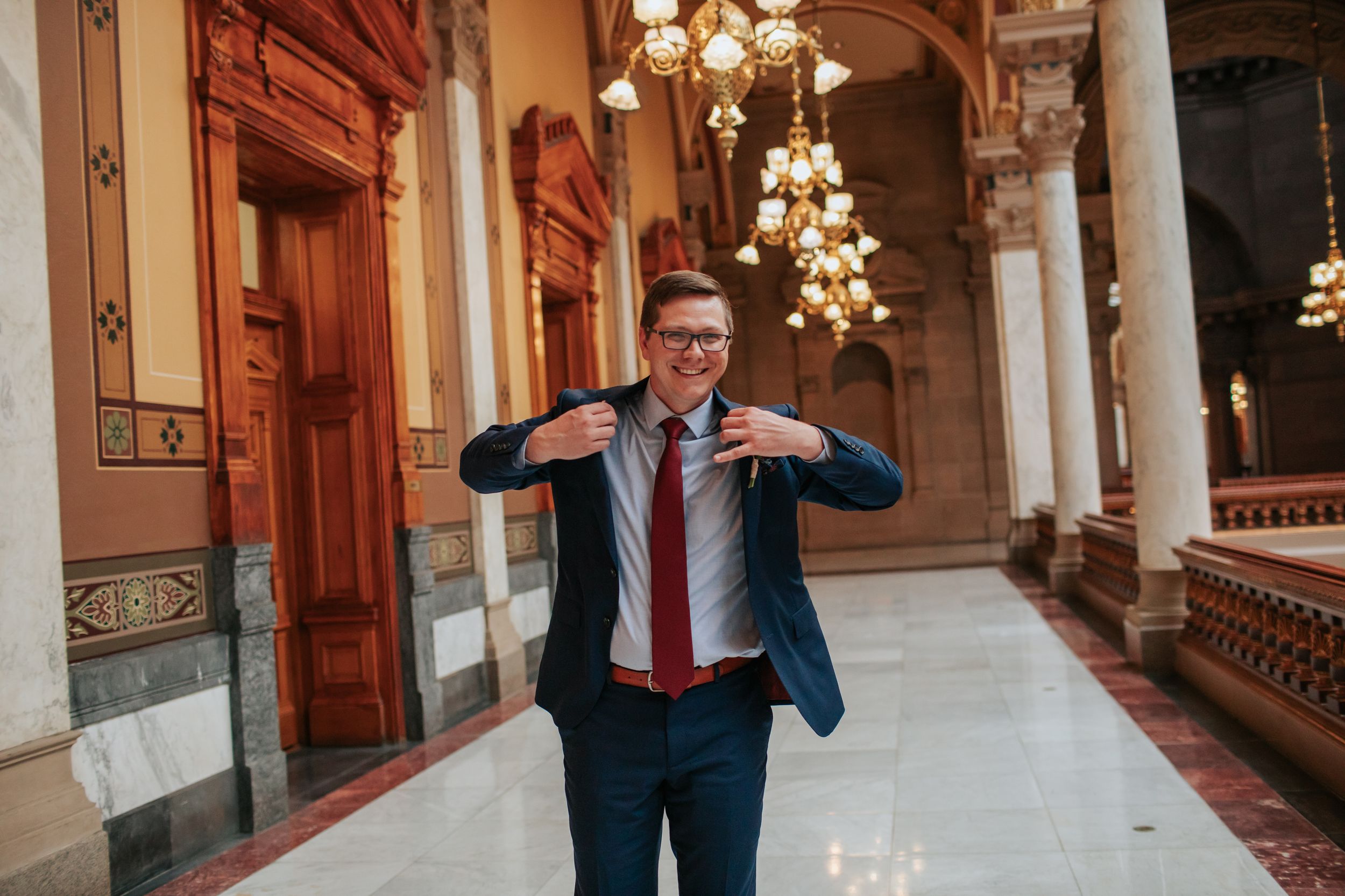 A person in a suit jacket poses playfully in an ornate hallway with classical architectural details and chandeliers.