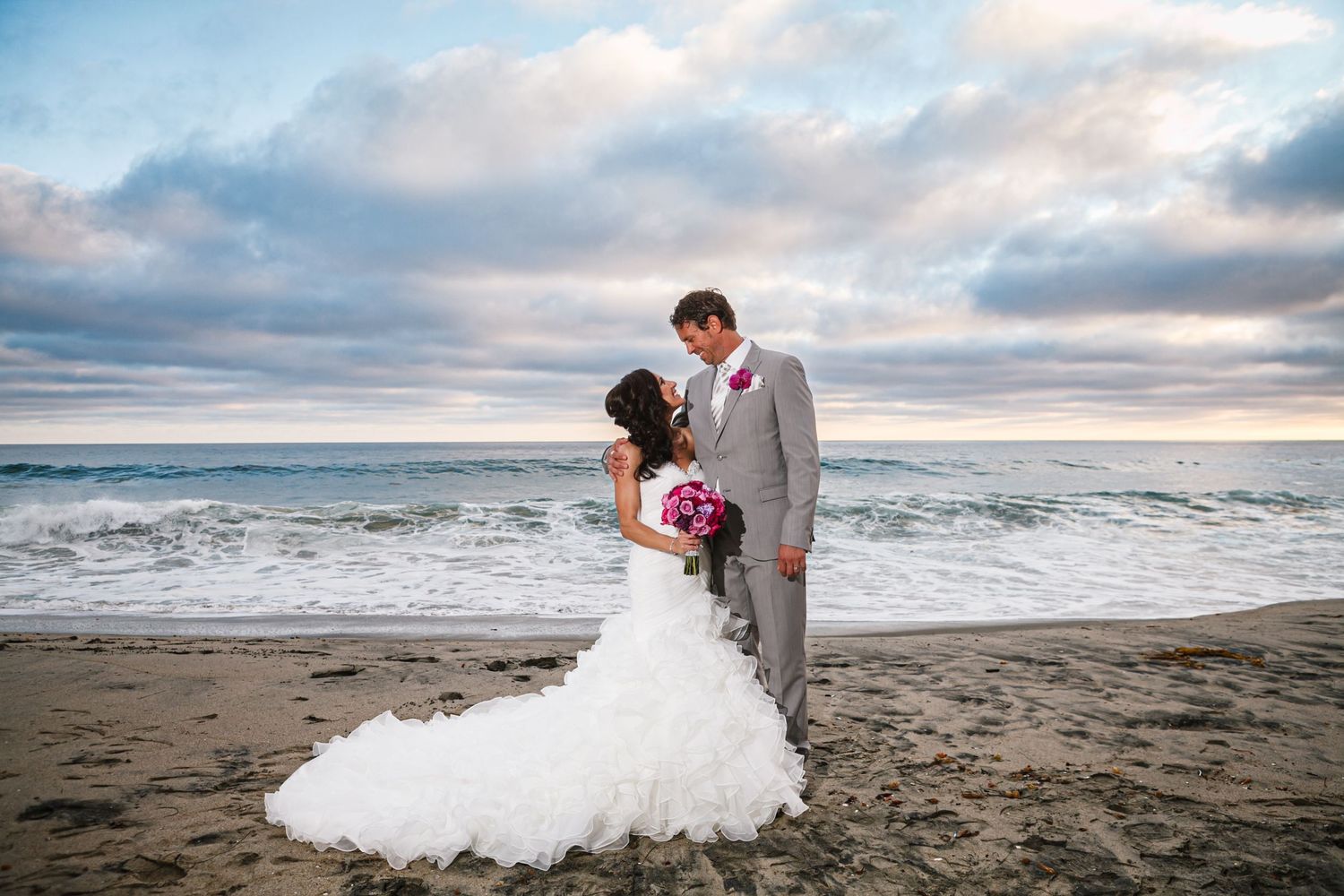 A couple in wedding attire stands together on a sandy beach with dramatic cloudy skies and ocean waves in the background.