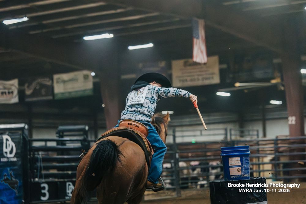 Indoor rodeo arena with handlers and riders preparing for competition under fluorescent lighting.
