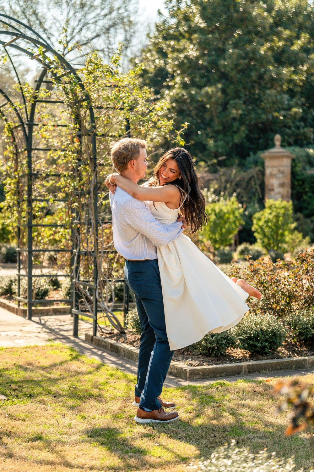 Man lifting woman in white dress in a sunlit garden with a metal arbor and lush greenery in the background.