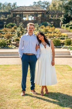 Couple posing in garden, man in blue pants and woman in white dress, with stone pergola backdrop.