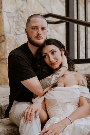 A couple poses intimately on stone steps, the woman in a white dress leaning on a tattooed man in a black shirt.