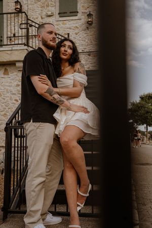 Couple posing on stone building steps, woman in white off-shoulder dress, man in black shirt with tattoos.
