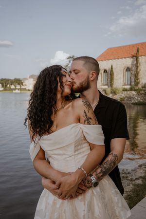 Couple sharing a romantic kiss in wedding attire by a lake, man with tattoos in black shirt embracing bride in off-shoulder gown.