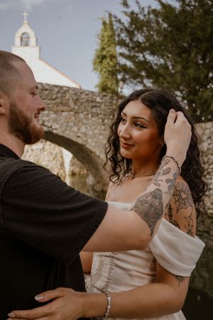 Tattooed bride in white dress gazes at bearded groom near stone arch and white chapel outdoors.