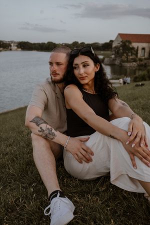 Couple sitting on grass by a lake at sunset, woman in black top with white skirt, man with tattoos in beige shirt.