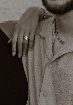 Close-up of a woman's hand with a diamond engagement ring resting on a man's shoulder, both wearing gold jewelry.