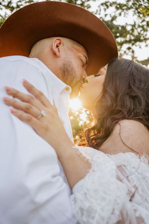 Couple sharing a romantic kiss at golden hour, man in brown cowboy hat and white shirt, woman in lace dress with engagement ring.