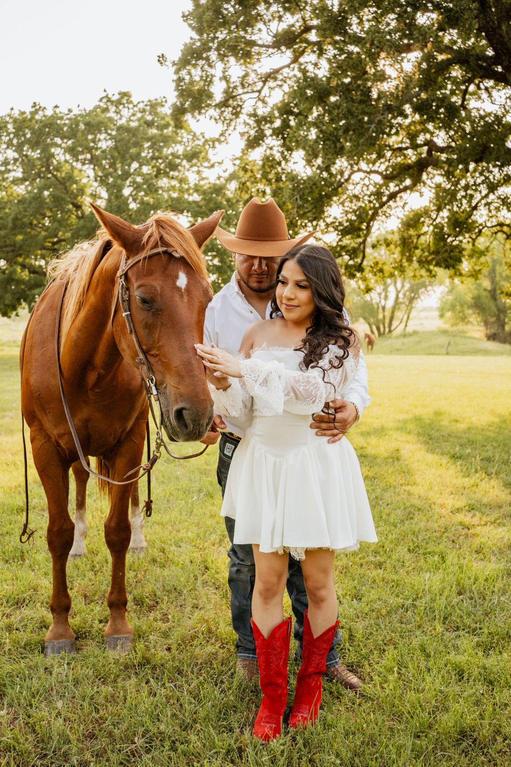 Couple in western attire with a chestnut horse outdoors, woman in white dress and red cowboy boots, man in brown hat.