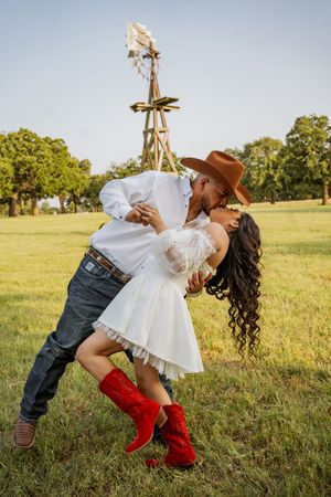 Couple sharing a romantic dip kiss on a green farm field, woman in white dress and red cowboy boots, man in brown hat.