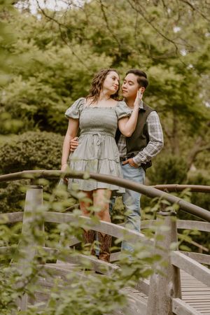 Couple sharing a romantic moment on a wooden bridge surrounded by lush green trees in a serene garden setting.