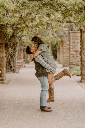 Man lifting woman in a joyful embrace on a tree-lined garden path, both wearing casual western-style outfits.