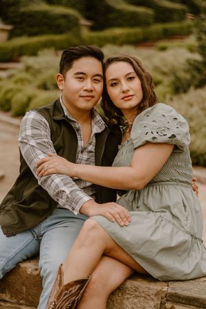 A young couple sits closely together outdoors, the man in a plaid shirt and vest, the woman in a sage green dress.