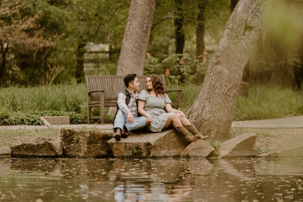 Couple sitting romantically by a serene pond in a lush green park, sharing a tender moment near large trees.