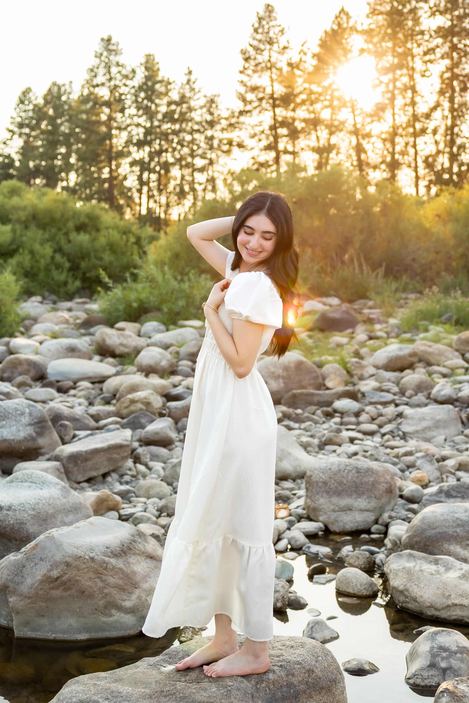 Ethereal figure in flowing white dress poses gracefully on rocky riverbed at golden sunset among tall pine trees.