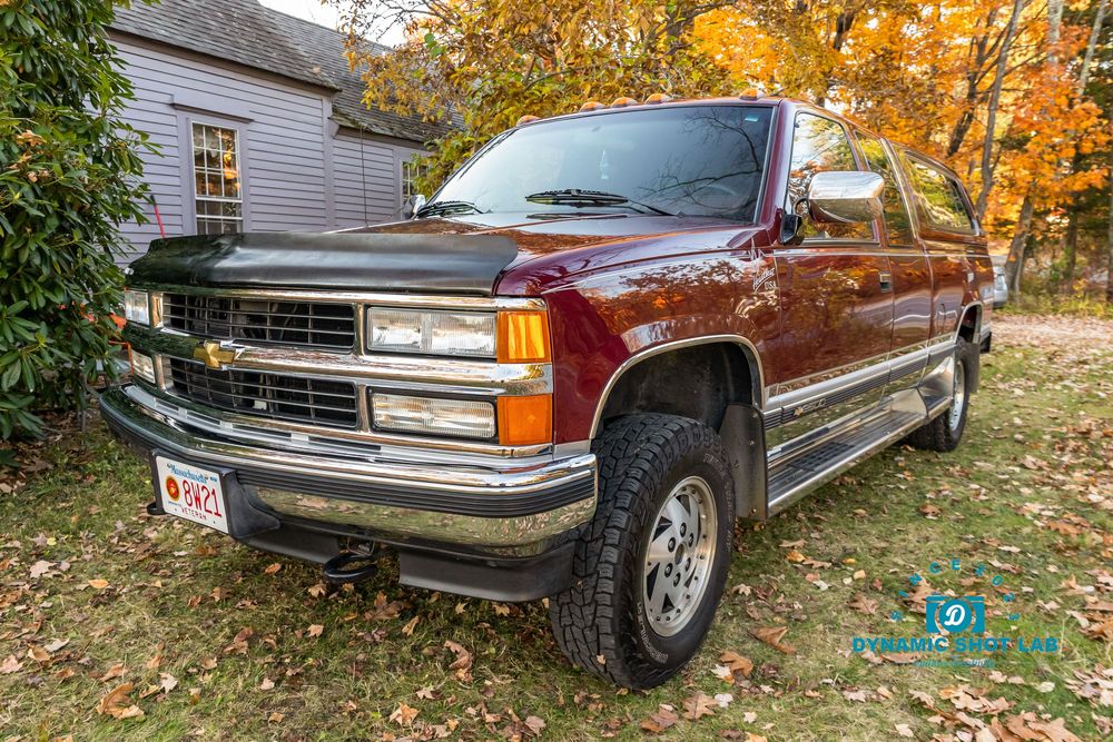 Red Chevrolet pickup truck parked outdoors with autumn foliage in background near a gray wooden building.