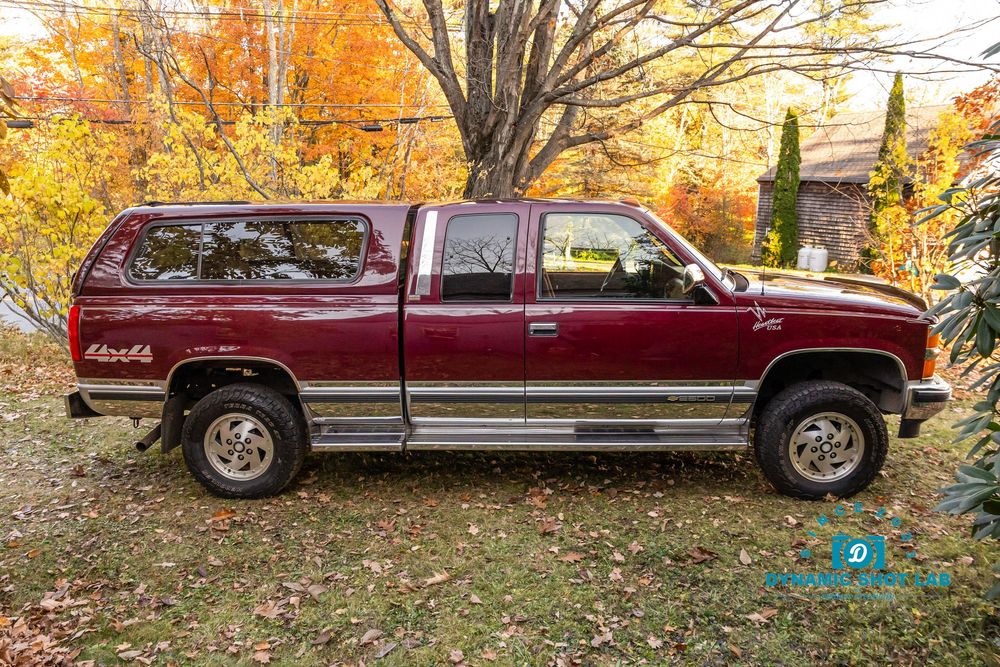 Maroon Chevrolet Silverado extended cab pickup truck with camper shell parked outdoors among fall foliage trees.