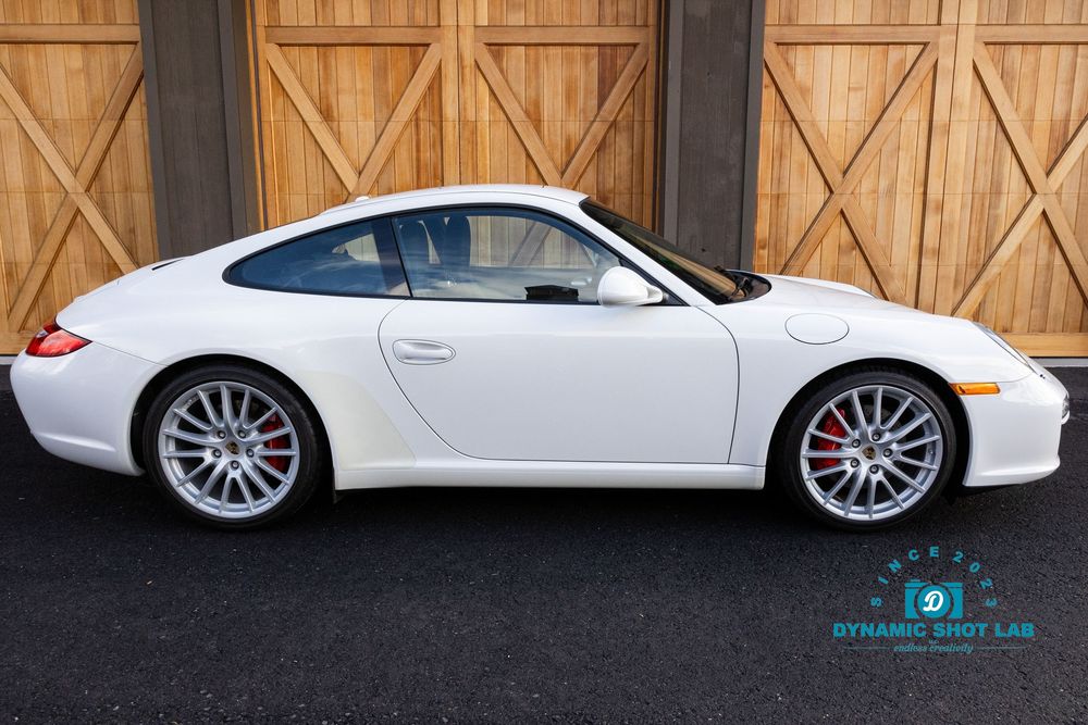 White Porsche 911 Carrera coupe parked in front of wooden garage doors, side profile view showcasing sleek design.