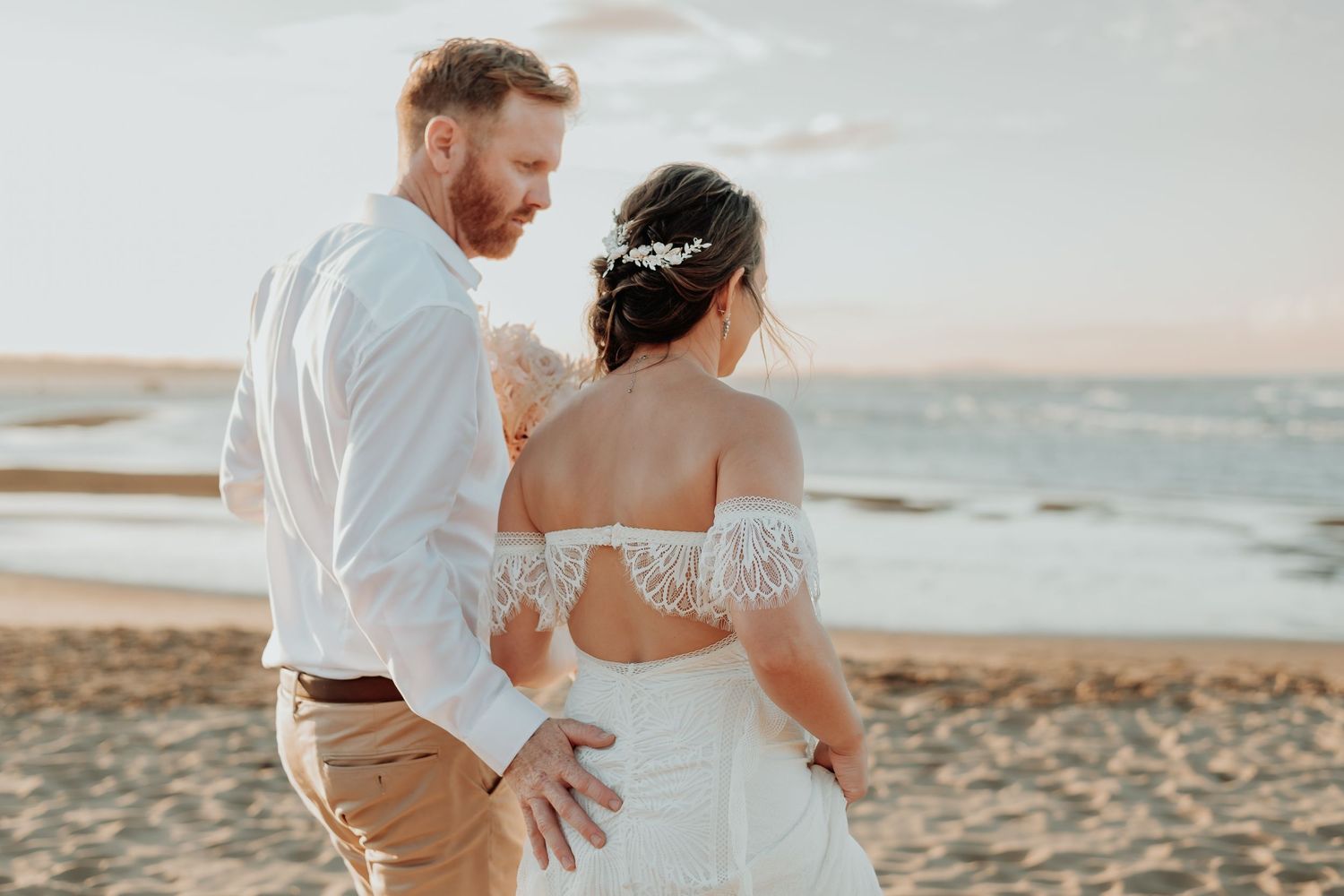 A couple embraces on a sandy beach at sunset wearing wedding attire during their romantic photoshoot.