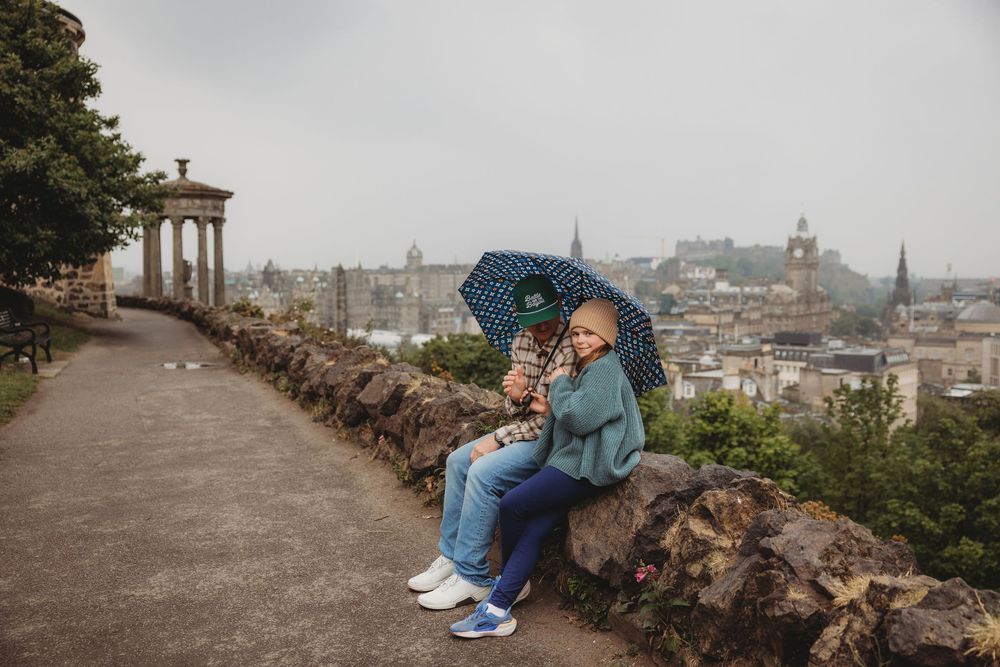 Panoramic view of Edinburgh city skyline with historic buildings and monuments visible through a stone wall walkway.