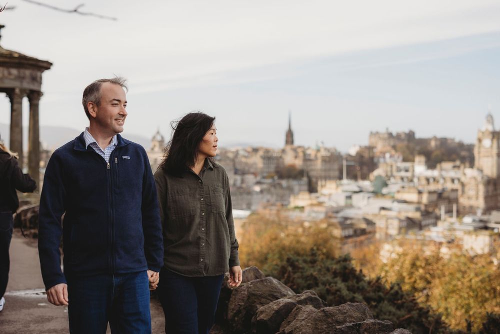 Two people stand on a rooftop terrace overlooking an autumn cityscape with historic buildings and fall foliage.