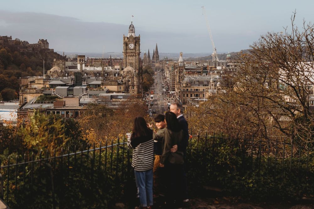 Two people admiring the Edinburgh cityscape with historic architecture and clock tower from a hilltop viewpoint.
