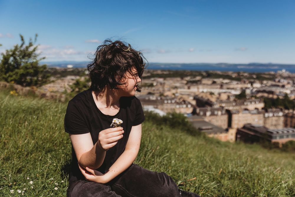 A romantic couple embraces on a grassy hilltop overlooking a scenic city panorama on a sunny day.