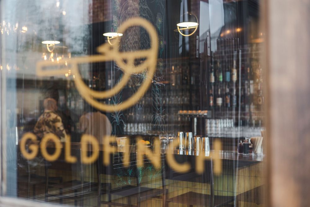 Modern cafe interior with large glass windows, wooden furniture and gold lettering signage visible from outside.