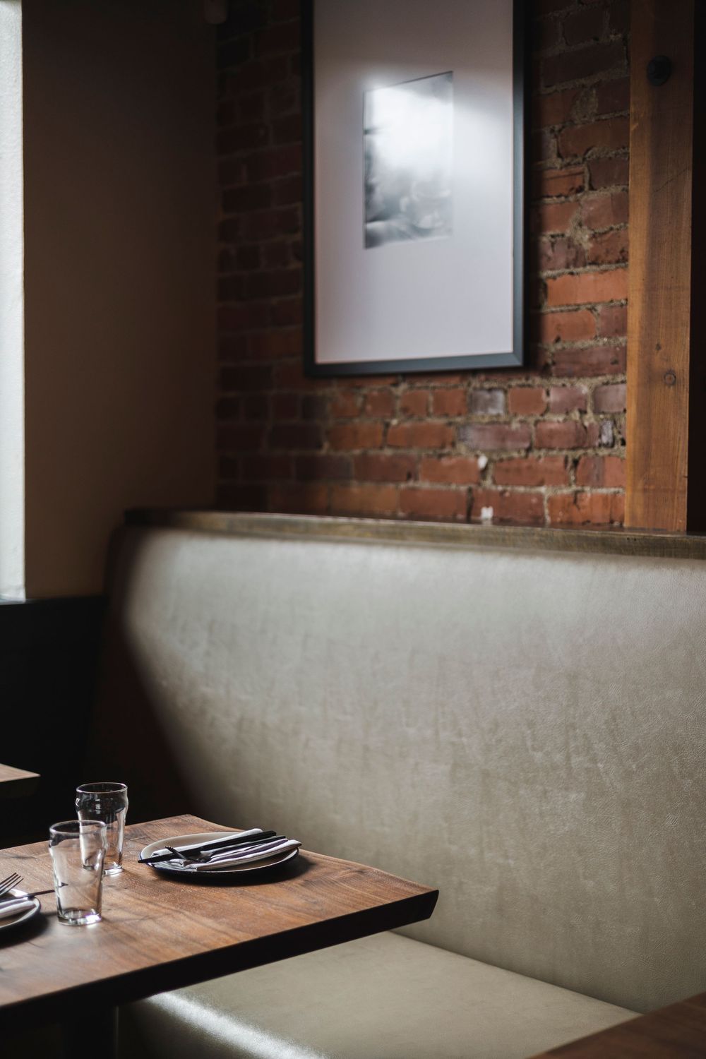 Modern restaurant interior with exposed brick walls, wooden tables, and grey banquette seating in moody lighting.