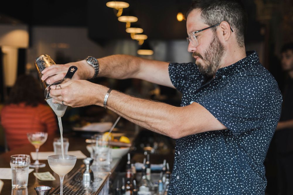 Bartender in a polka dot shirt pours multiple cocktails simultaneously at a dimly lit bar counter.