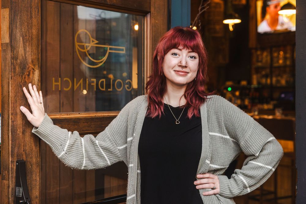Person with red hair wearing a gray cardigan stands in front of a wooden wall with warm lighting in the background.