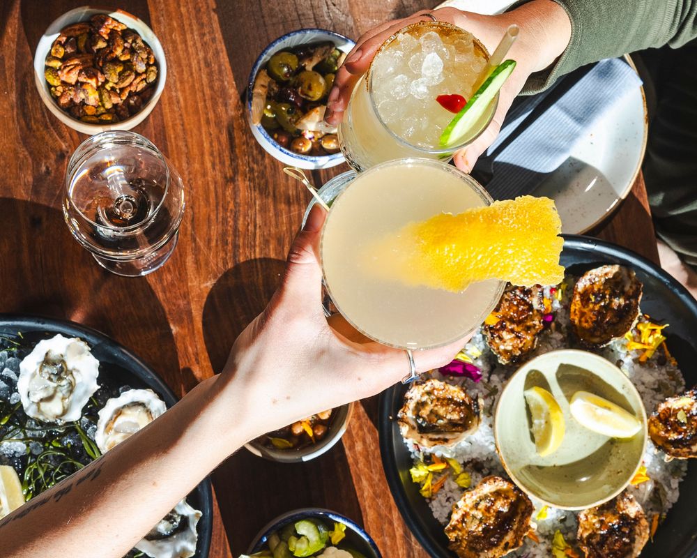 Hands holding cocktail glasses with citrus garnishes over a dark wooden table with scattered drinks and snacks.