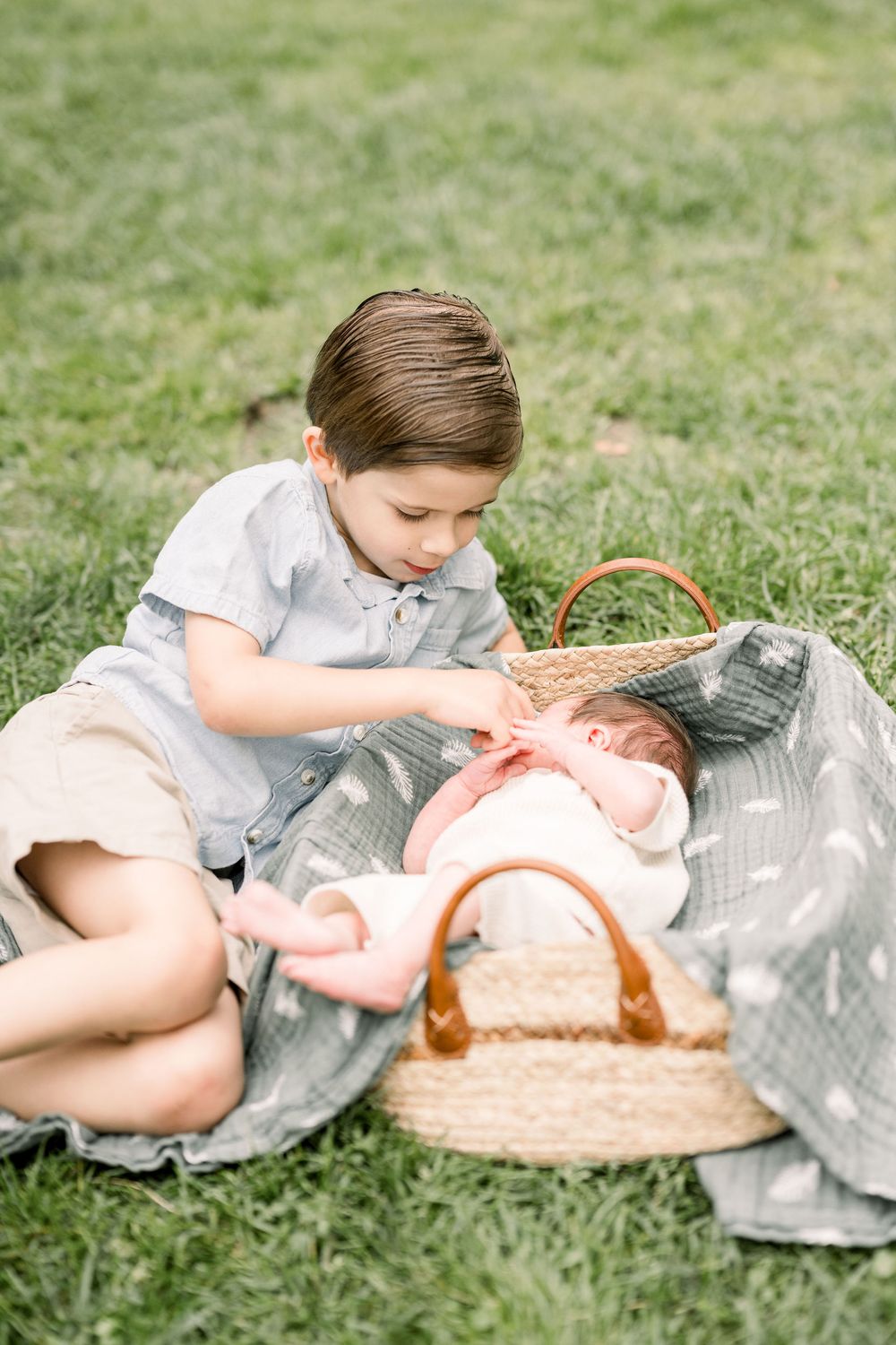 Child in white shirt and gray pants lying on blanket reaching for wicker basket during outdoor picnic.