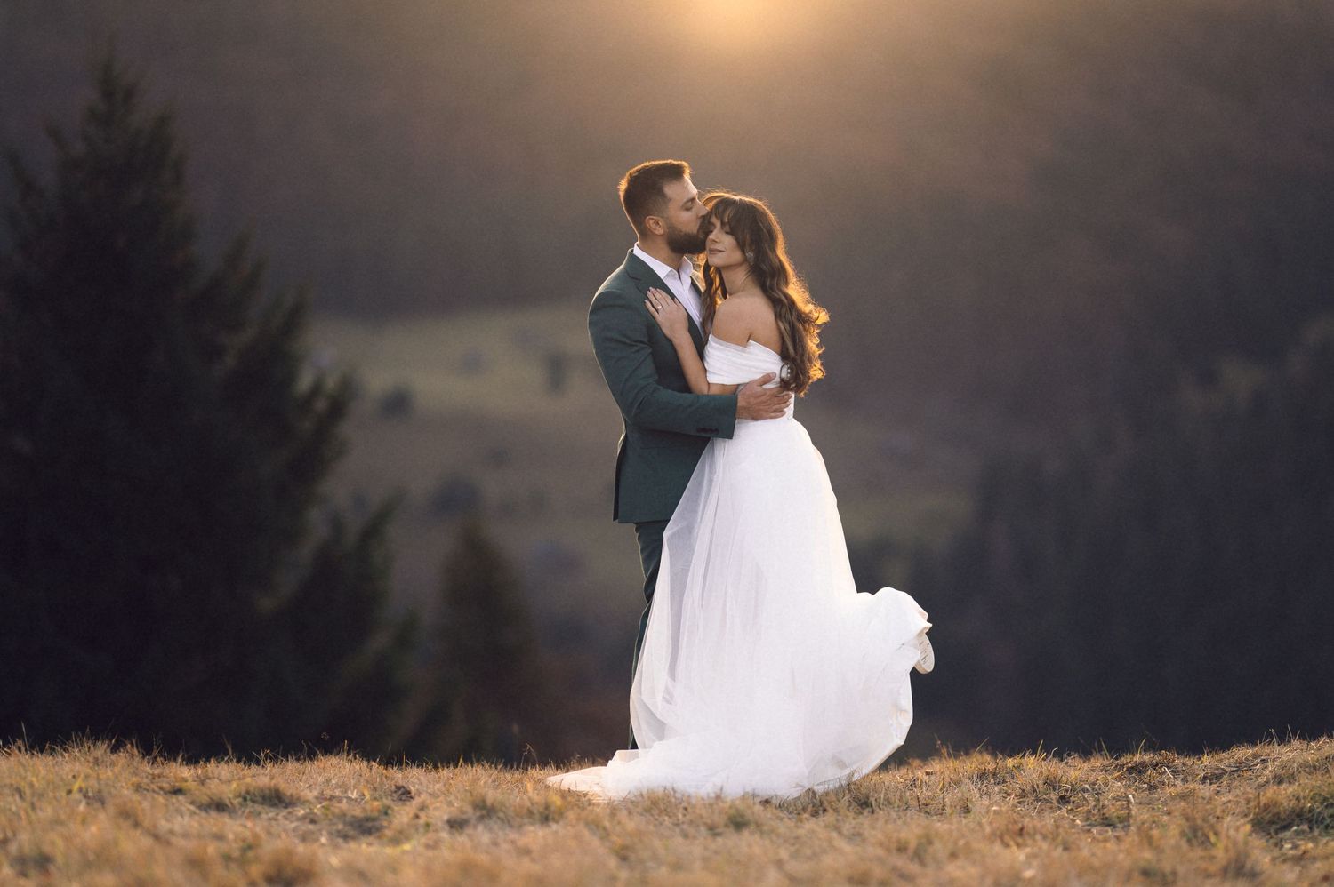 A couple in wedding attire embraces on a hilltop against a dramatic sunset sky.