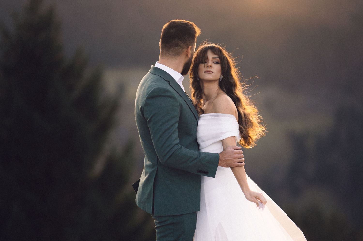 A couple in wedding attire embrace during sunset, with the bride in white and groom in dark green suit.