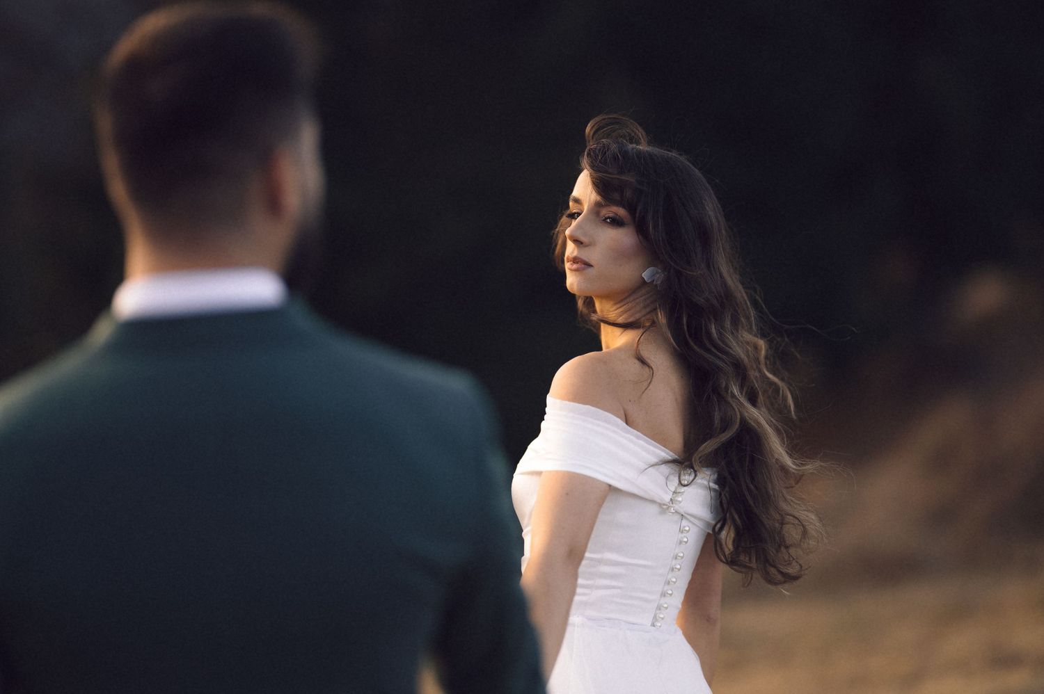 A bride in a white wedding dress poses against a dark background during an outdoor photo session.