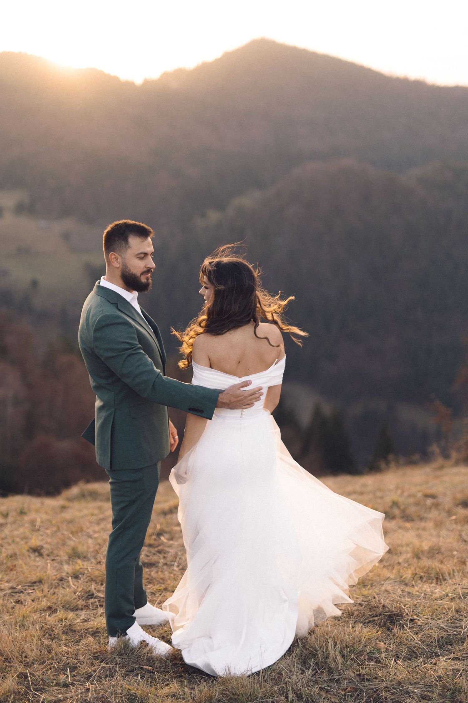 A couple dances on a hilltop at golden hour, with mountains in background and bride's dress flowing in the wind.
