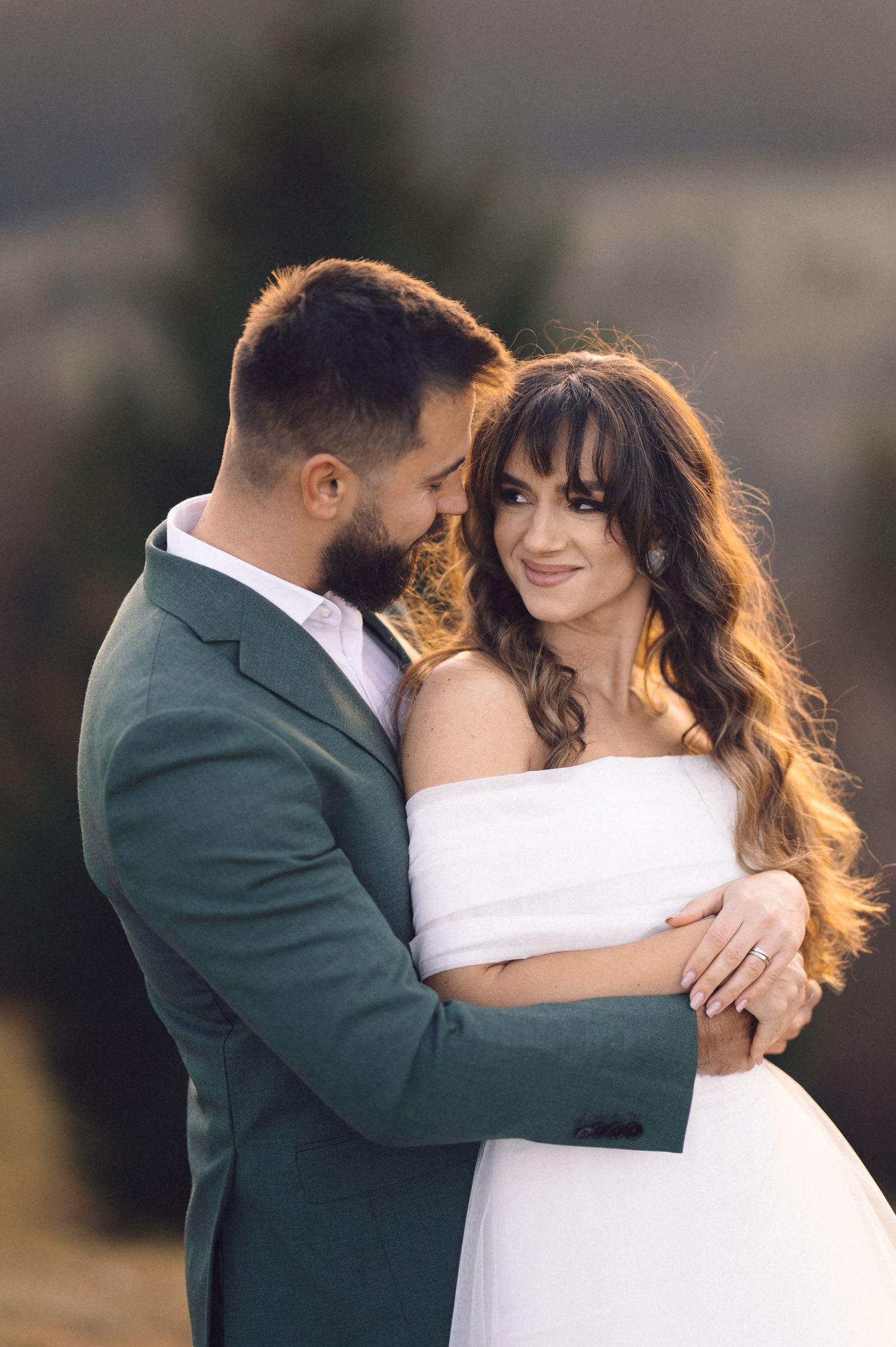 A romantic wedding portrait of a couple embracing at sunset, with the bride in white and groom in green suit.