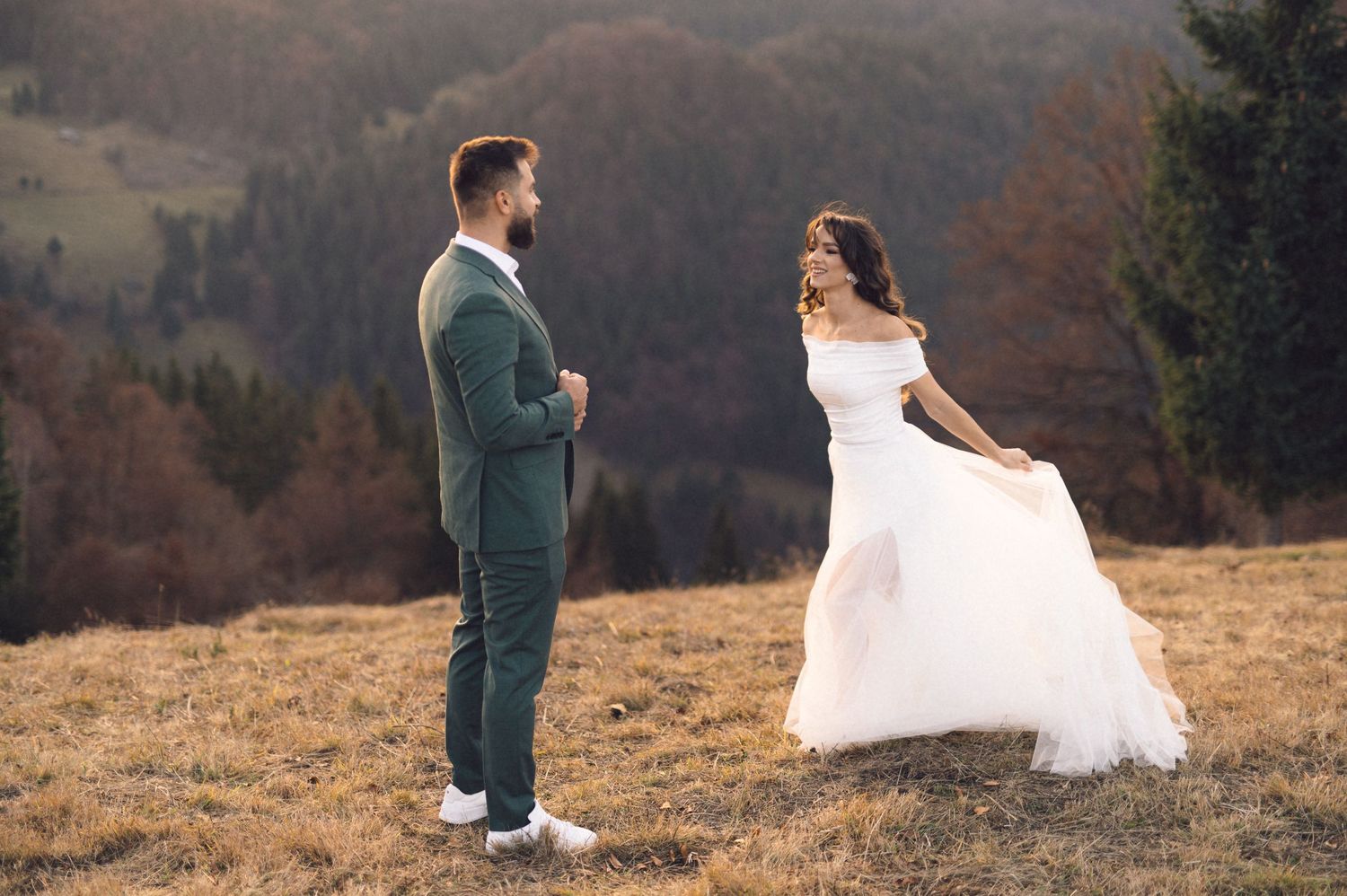 A couple in wedding attire stands together on a scenic mountain hillside during an autumn sunset.