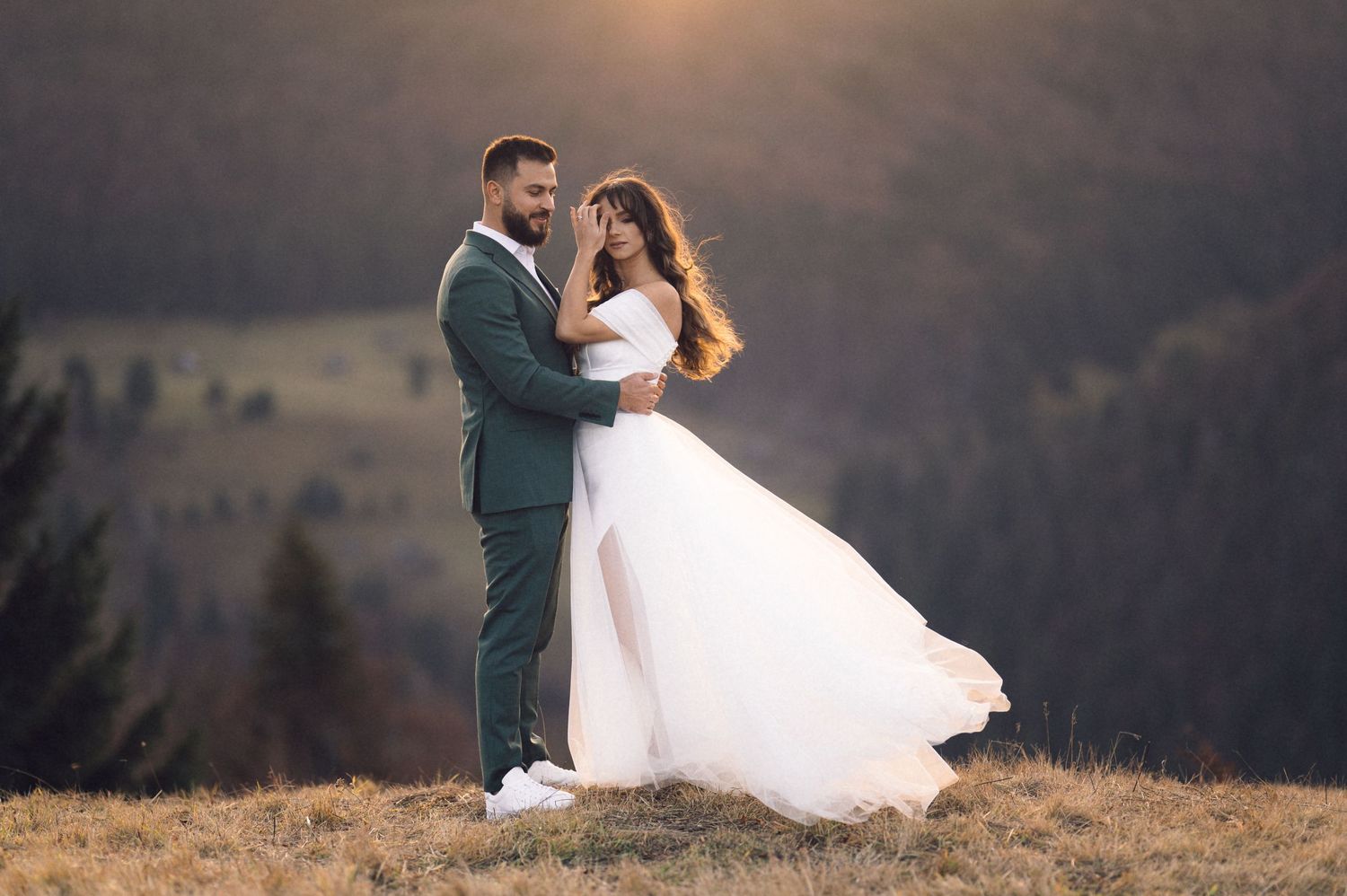 Newlywed couple embraces during sunset photoshoot in scenic mountain landscape with flowing white wedding dress.