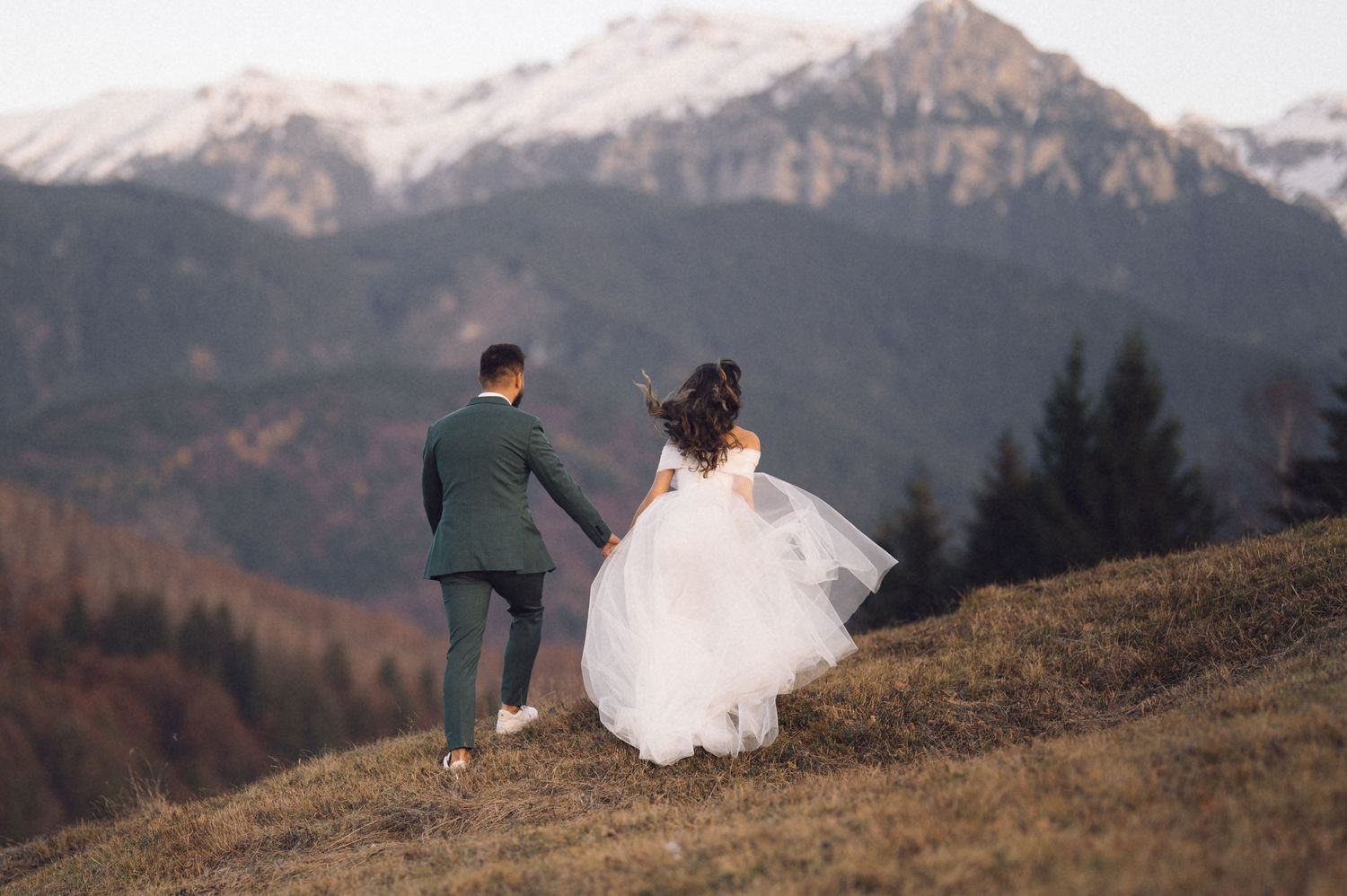 A couple in wedding attire stands on a mountain ridge overlooking snow-capped peaks during sunset.