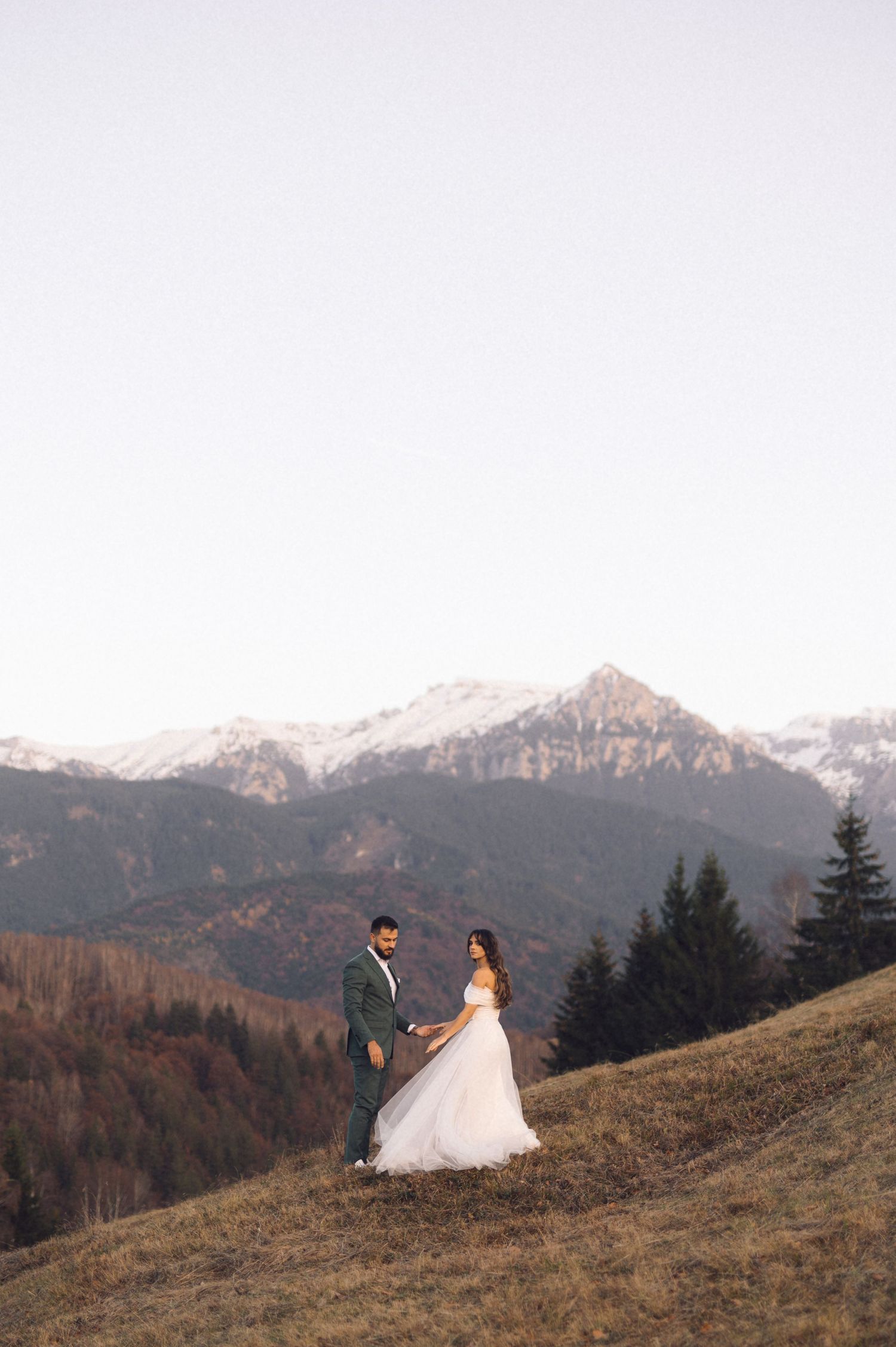A couple in wedding attire stands on a mountainside slope with snow-capped peaks and evergreen forest in the background.