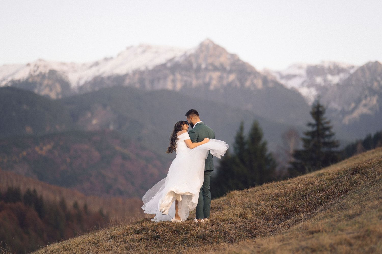 A couple in wedding attire embraces on a mountainside with snow-capped peaks in the background at sunset.