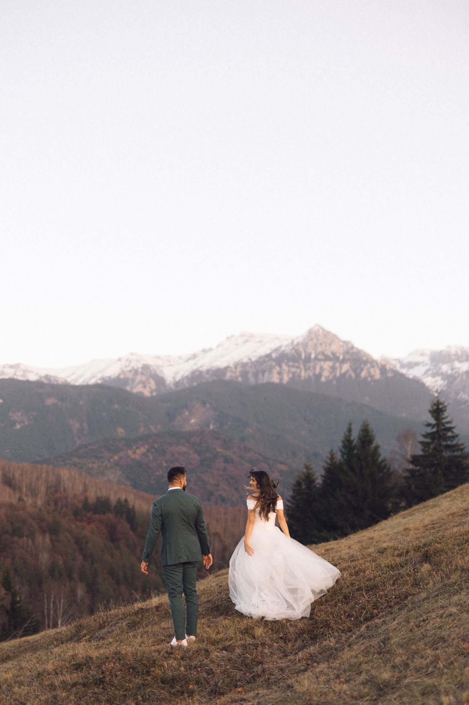A couple stands on a scenic mountain slope with snow-capped peaks in the distance during golden hour.