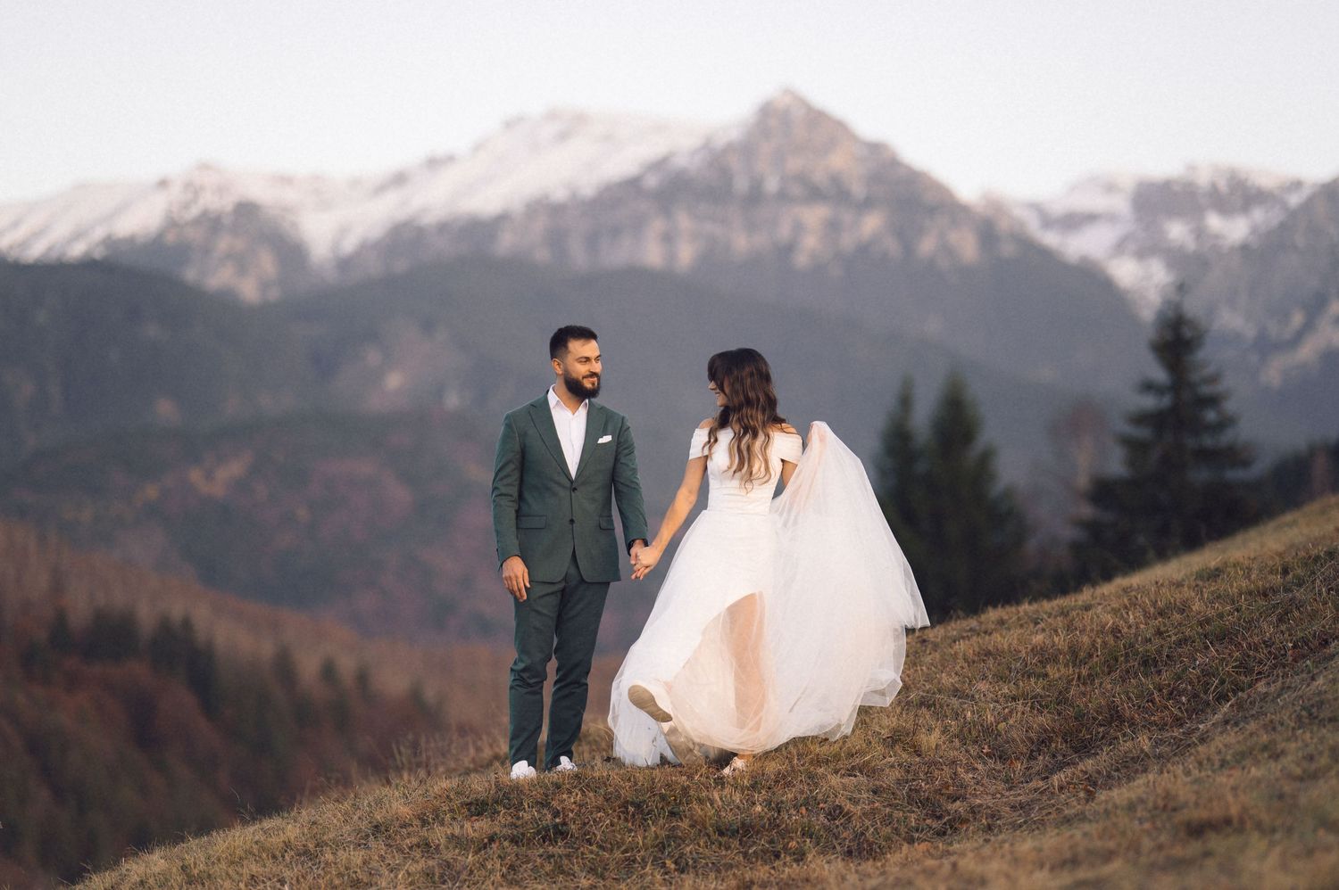A couple in wedding attire poses on a mountain ridge with snow-capped peaks and evergreen forest in the background.